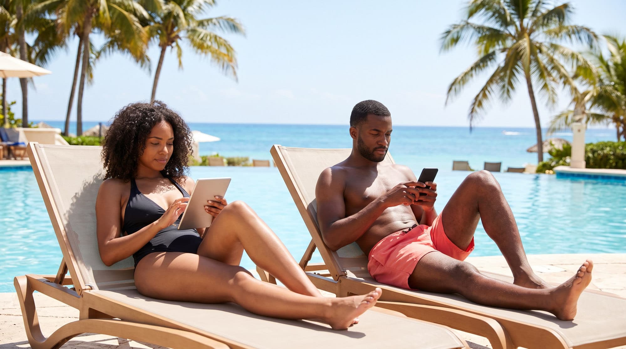 A couple on sun loungers beside a hotel pool, one reading a tablet and the other using a phone.