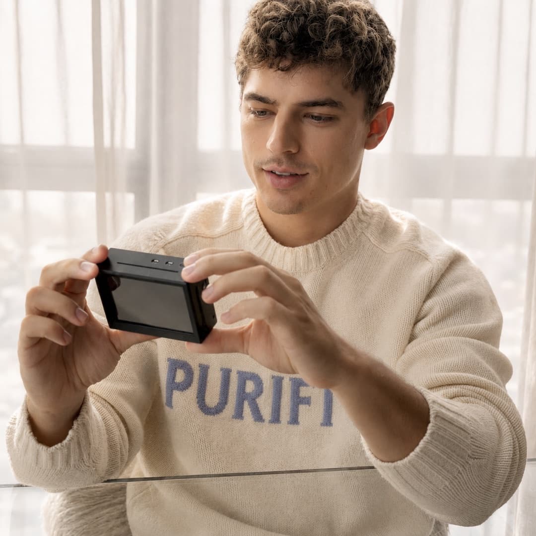 A man holding the small black PuriFi device in both hands beside a sunlit window.